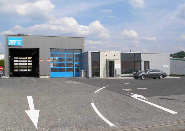 Ein modernes Gebäude mit einem blauen Garagentor, einem weißen Schild mit der Aufschrift "TÜV" und einem grauen Auto, das davor geparkt ist.