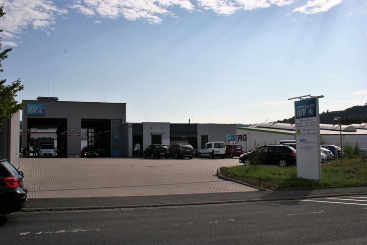 Eine moderne Autowerkstatt mit einem Schild mit der Aufschrift "TUEV Hessen" und einer Liste von Dienstleistungen. Draußen sind mehrere Autos geparkt, und der Himmel ist klar mit ein paar Wolken.