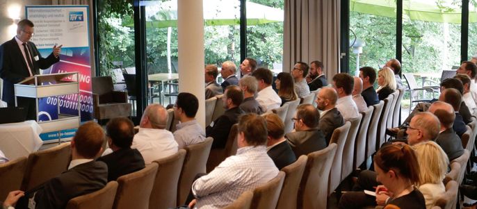 Ein Raum voller Menschen, die in Reihen sitzen und einem Redner auf dem Podium aufmerksam zuhoeren.