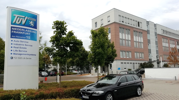 Ein großer Pylon von "TUEV Hessen" mit verschiedenen Dienstleistungen, darunter "Auto-Service", "Industrie-Service" und "Immobilien". Im Hintergrund steht ein modernes Gebäude mit mehreren Fenstern.